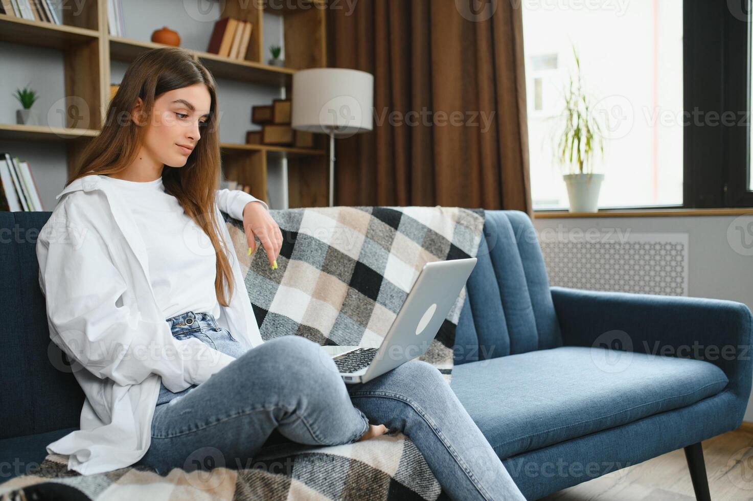 sorridente atraente jovem mulher sentado em sofá usando computador portátil comunicando trabalhando conectados às lar, feliz adolescente menina digitando em computador, desfrutando escrevendo blog ou conversando com amigos dentro social rede foto