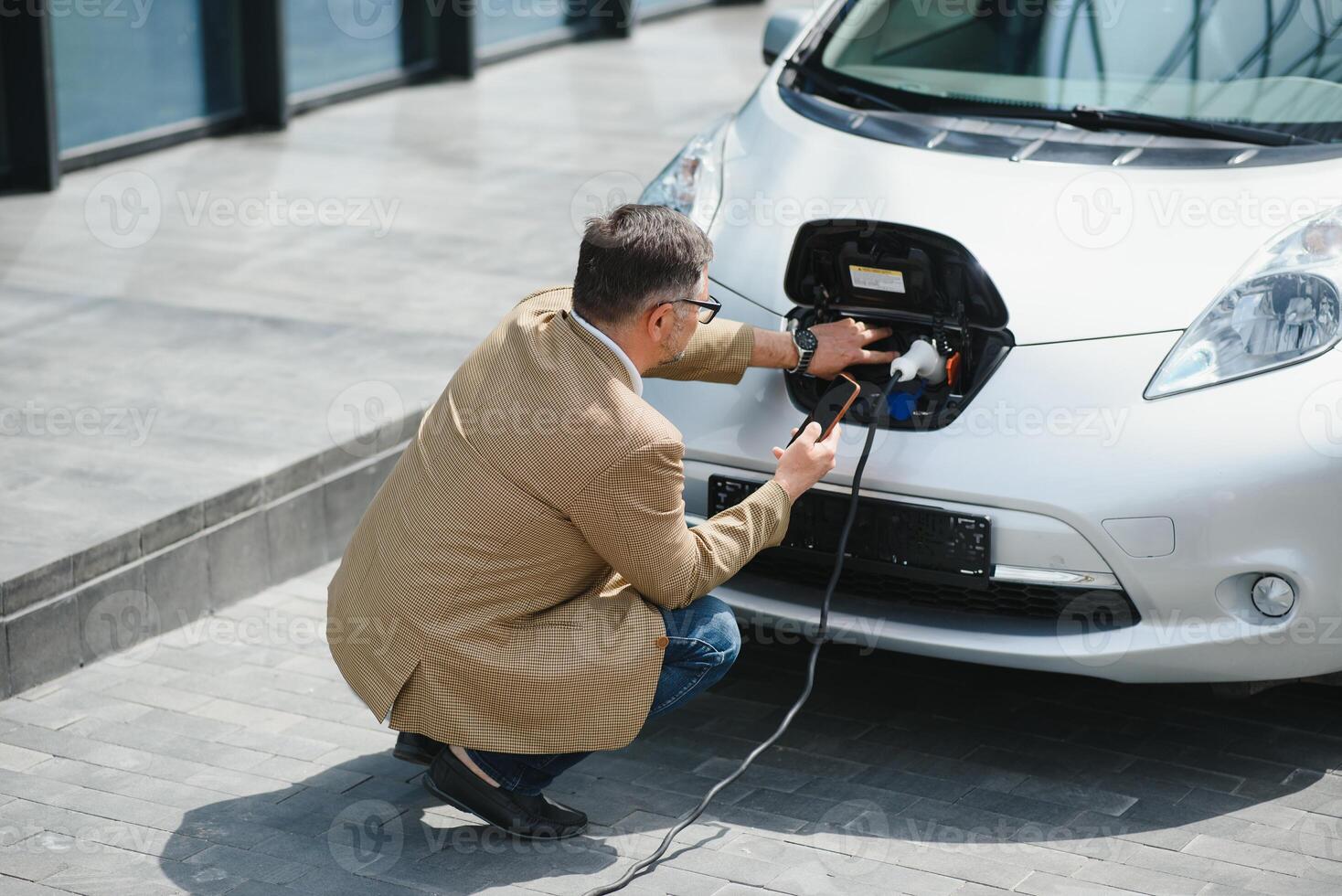 hansome barbudo homem sentado perto dele Novo moderno elétrico carro e segurando plugue do a carregador, enquanto carro é cobrando às a cobrando estação foto