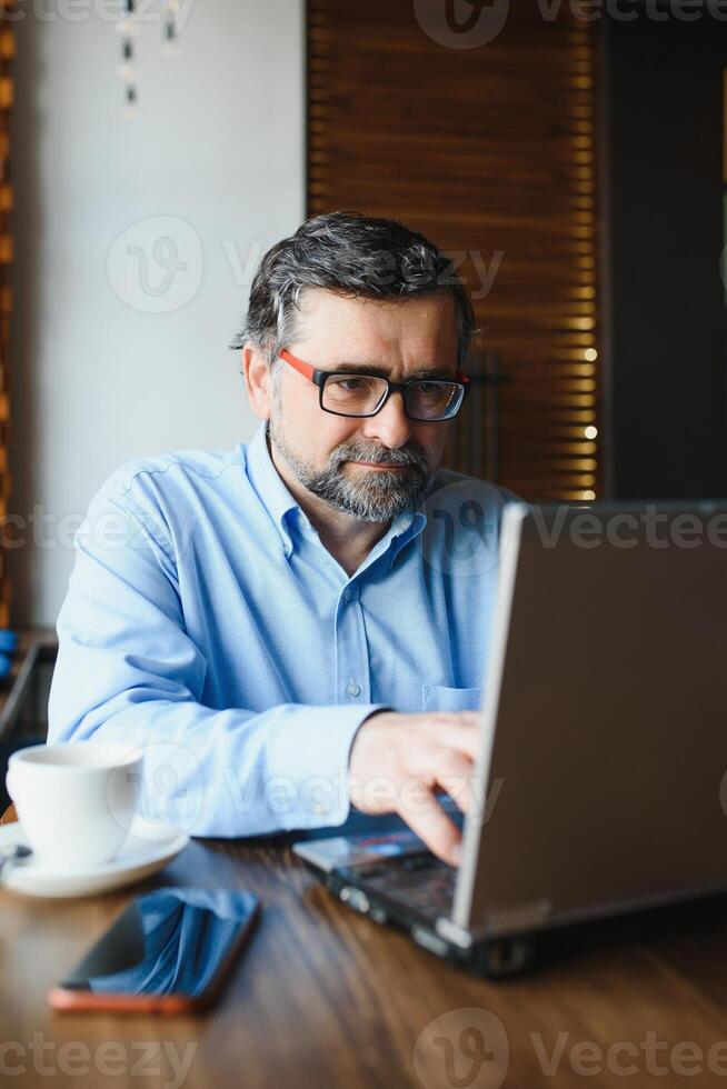 positivo Senior barbudo homem com cinzento cabelo bebendo café e usando computador portátil às cafeteria, cópia de espaço. à moda envelhecido homem de negocios dentro Borgonha Jaqueta desfrutando dele chá enquanto trabalhando on-line. foto