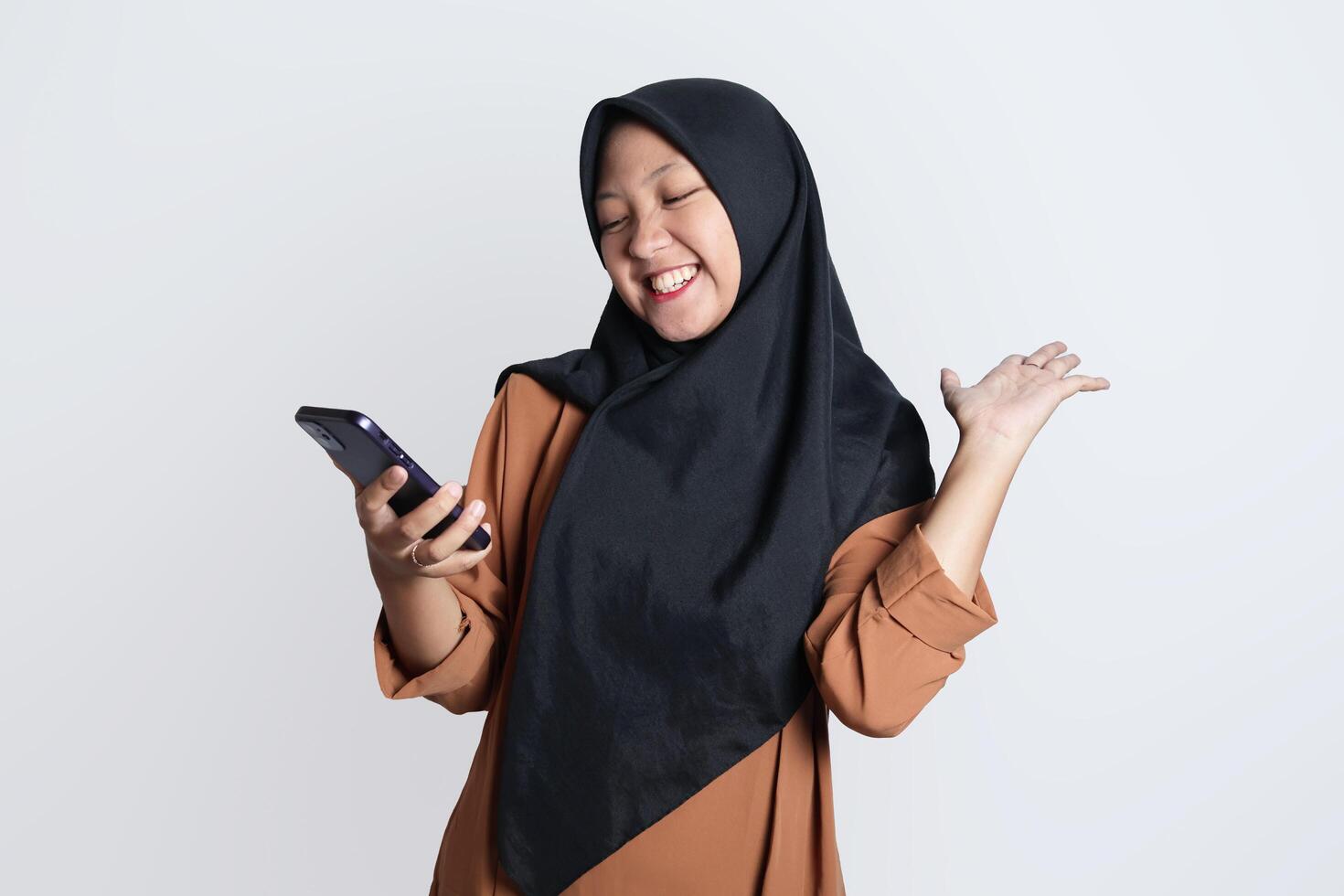 animado lindo ásia mulher dentro Castanho camisa e hijab usando Móvel telefone, a comemorar sucesso, obtendo Boa notícia isolado em branco fundo foto