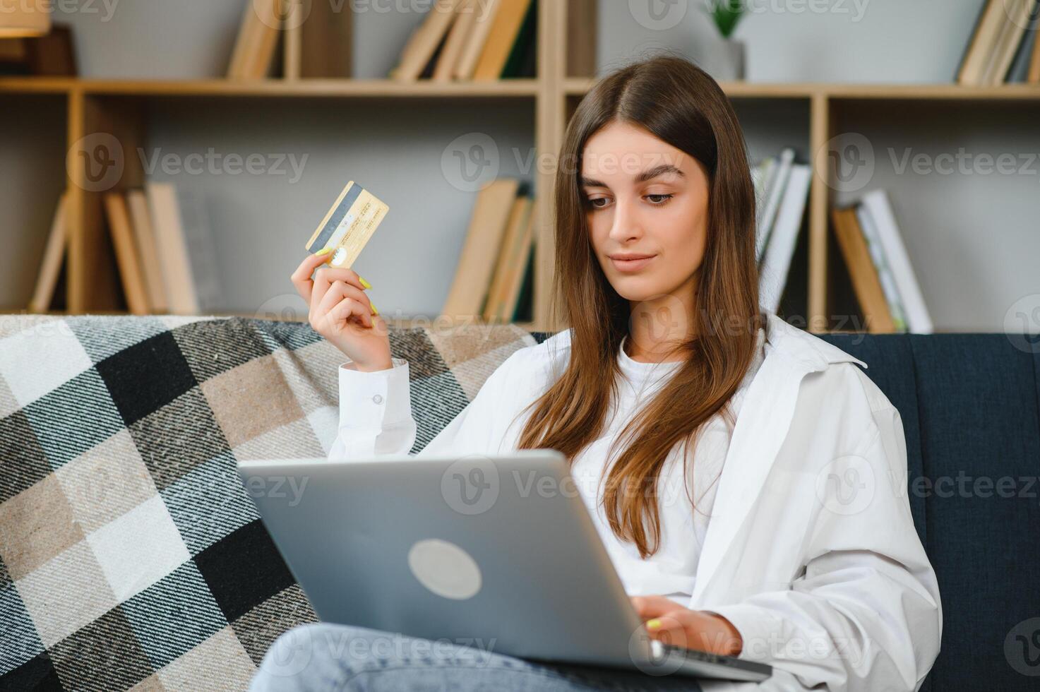 sorridente mulher segurando crédito cartão e usando computador portátil em sofá dentro vivo sala. foto