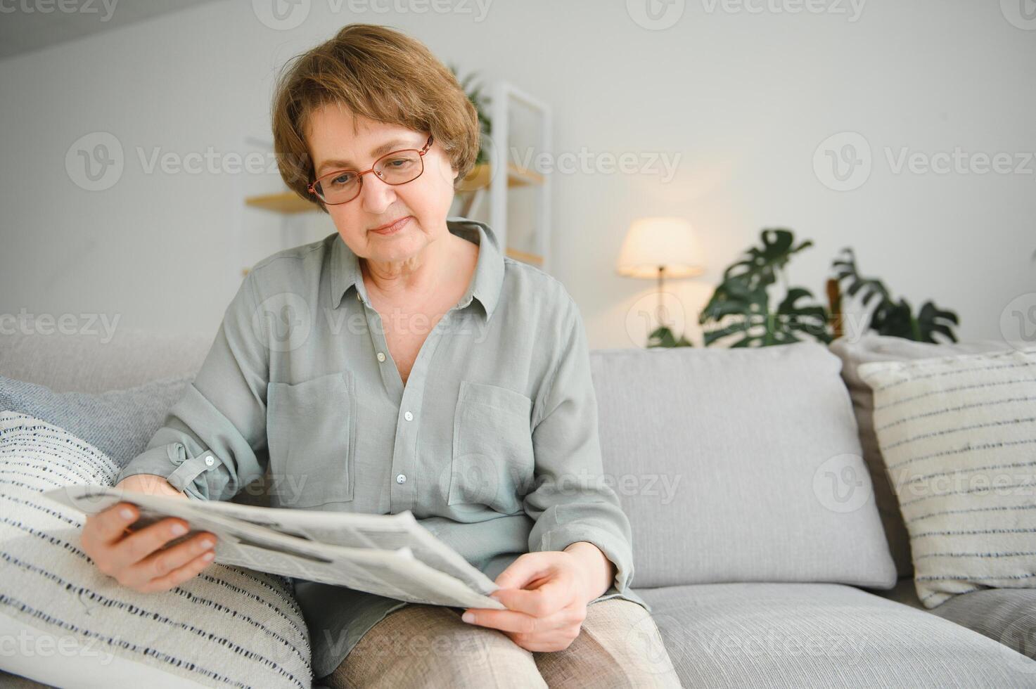 Senior senhora lendo dela jornal às casa relaxante em uma sofá e encarando sobre a topo às a espectador foto