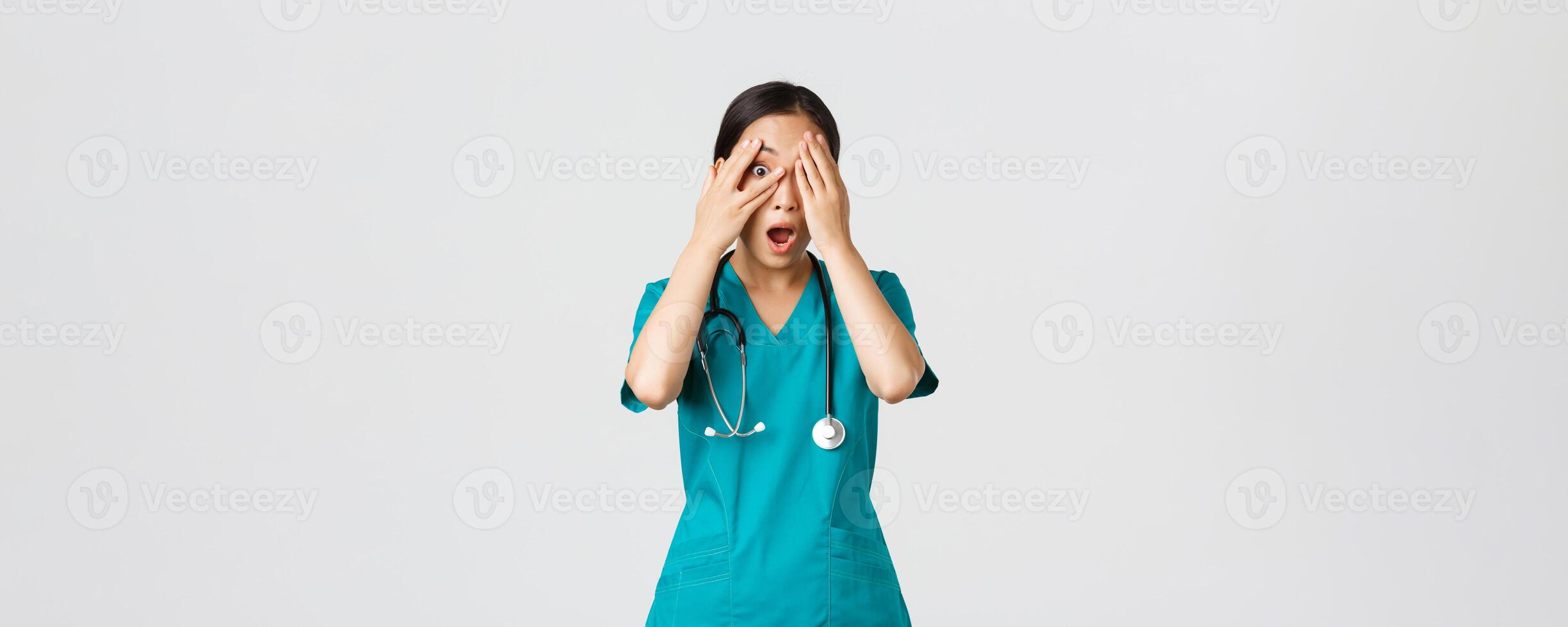 COVID-19, cuidados de saúde trabalhadores e impedindo vírus conceito. assustado e chocado ásia fêmea enfermeira, médico dentro esfrega testemunha alguma coisa embaraçoso ou apavorante, cobrir olhos mas olhadinha através dedos foto