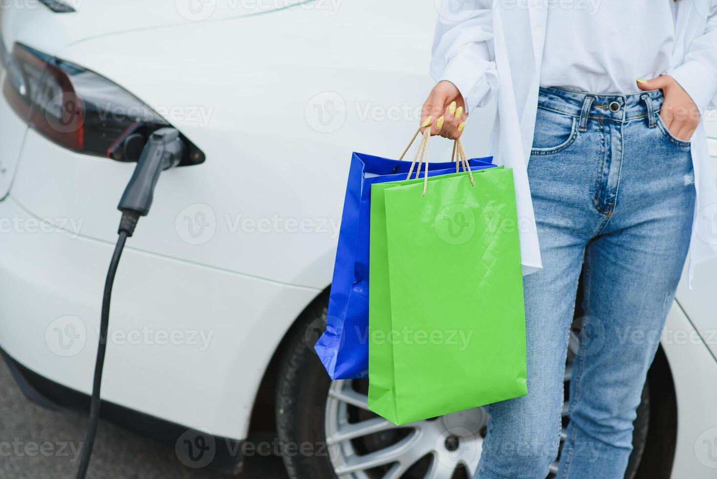 feliz jovem mulher em pé em cidade estacionamento perto elétrico carro, cobrando automóvel bateria a partir de pequeno cidade estação, segurando compras bolsas. foto