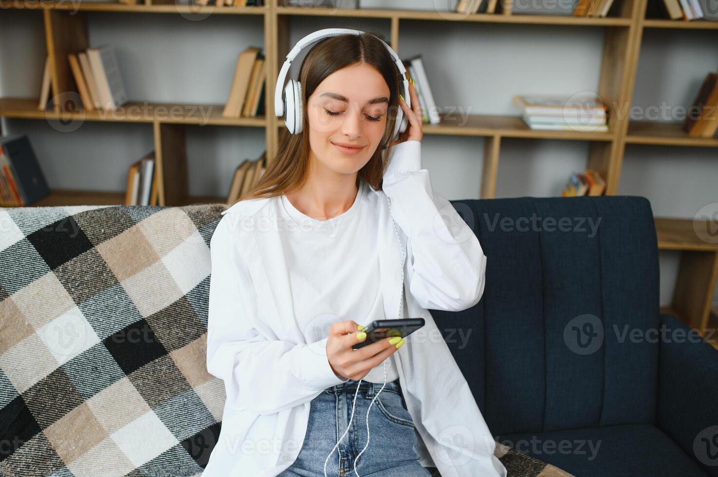 retrato do atraente menina 20s vestindo fones de ouvido usando Smartphone e ouvindo para música enquanto sentado em sofá às casa foto