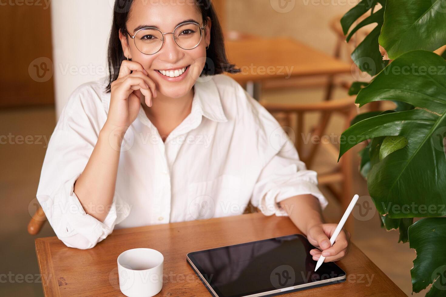 jovem ásia criativo garota, sentado com digital tábua, desenhando com gráfico caneta e sorridente, rabiscar, bebendo café dentro cafeteria foto