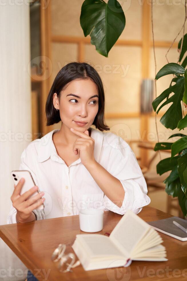 lindo jovem mulher, 25 anos velho, pensamento, segurando Smartphone e olhando considerado, sentado dentro cafeteria, decidir smth foto