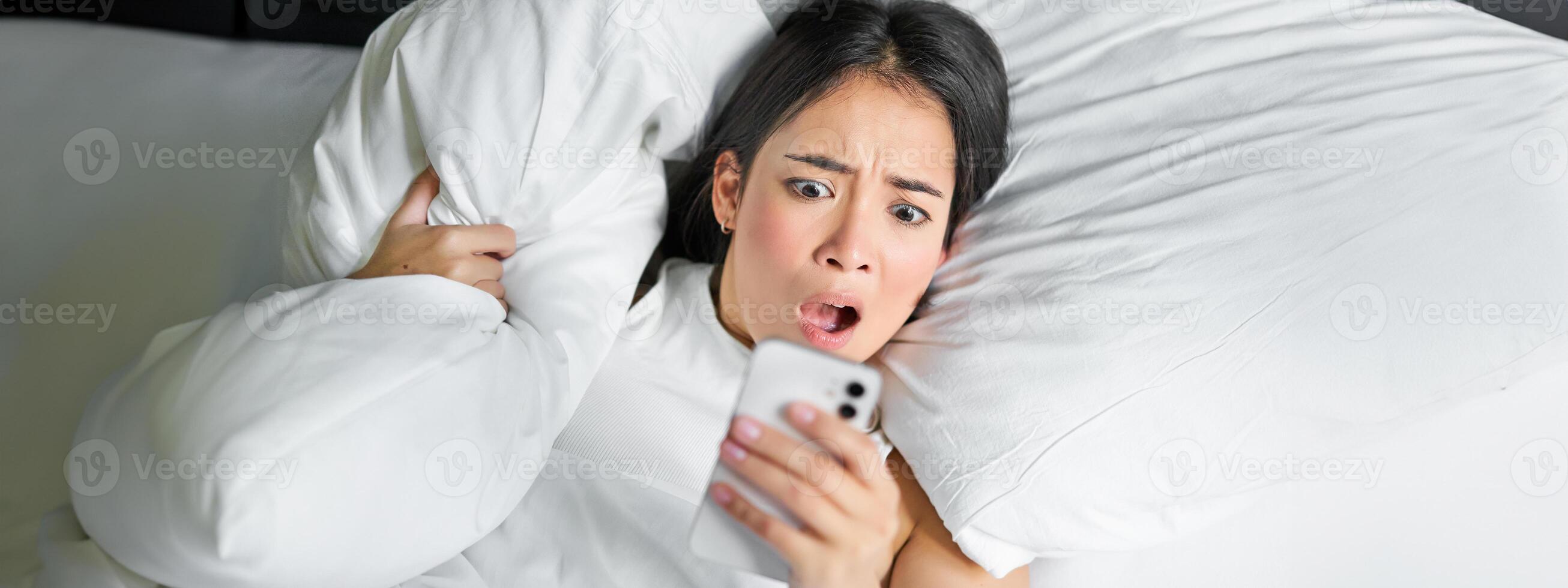 fechar acima retrato do ásia menina deitado dentro cama, olhando às Smartphone preocupado, acordando acima atrasado e encarando às dela alarme relógio em Móvel telefone foto