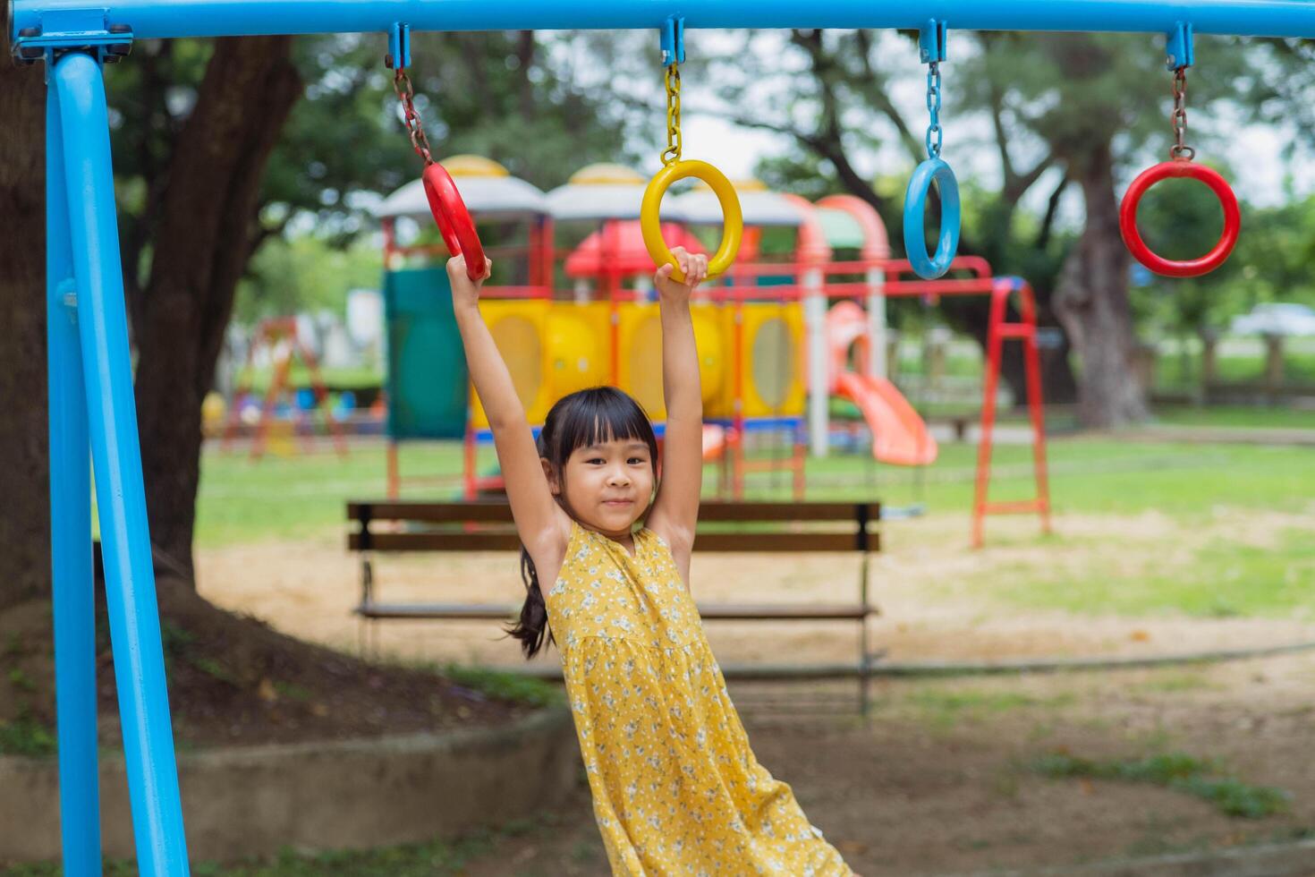 feliz menina suspensão em macaco Barra de mão fazendo exercício. pequeno ásia menina jogando às ao ar livre Parque infantil dentro a parque em verão período de férias. saudável atividade. foto