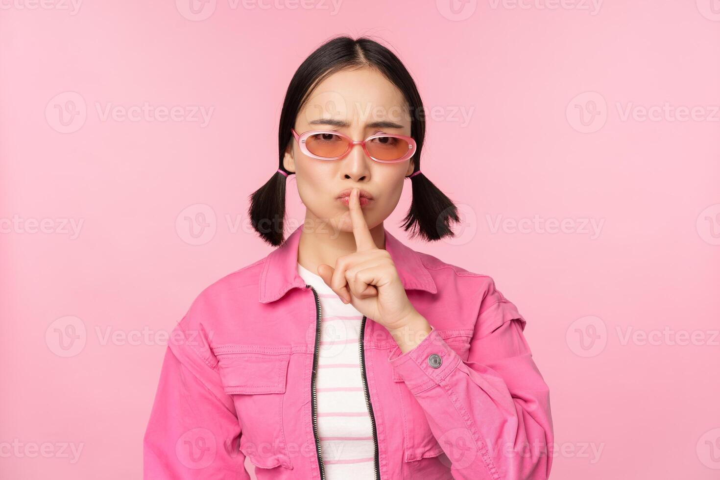 moderno coreano menina dentro à moda Primavera roupa, oculos de sol, mostrando cale-se, silêncio sinal, pressione dedo para lábios, tabu gesto, em pé sobre Rosa fundo foto