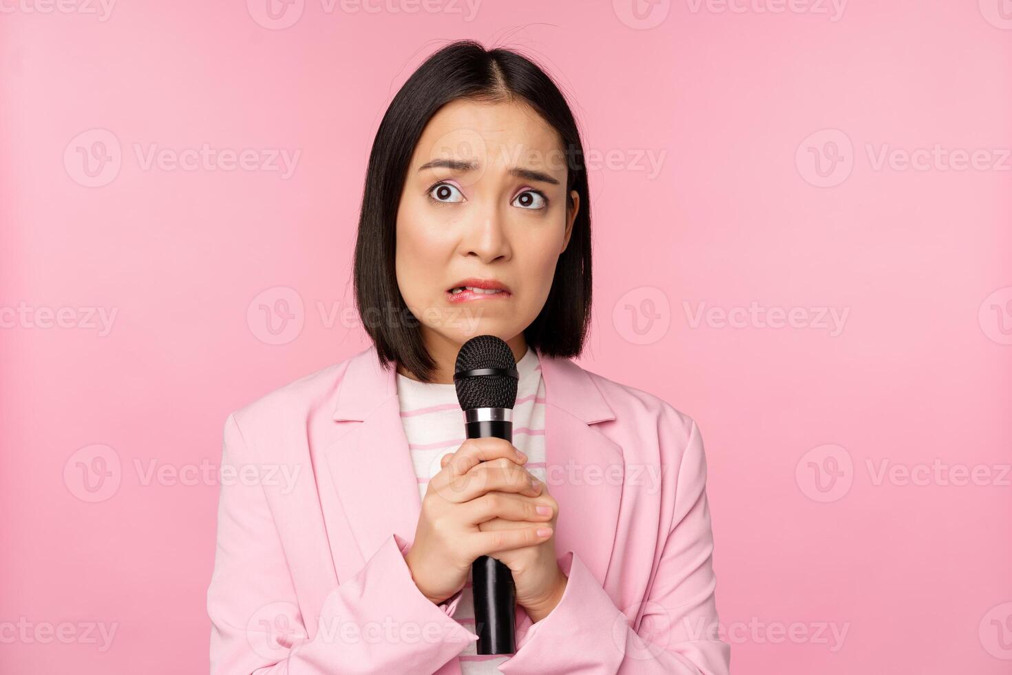 ansioso ásia senhora dentro terno, falando dentro público, dando discurso com microfone em conferência, olhando assustado, em pé sobre Rosa fundo foto