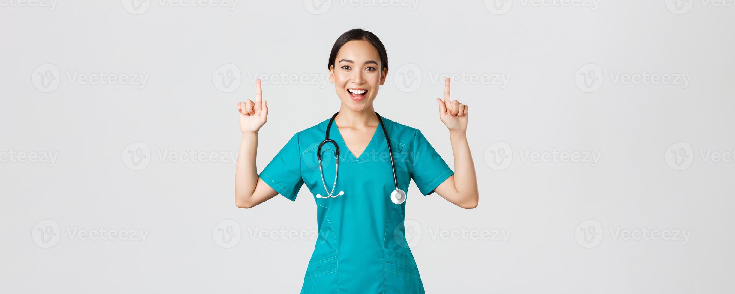 COVID-19, cuidados de saúde trabalhadores, pandemia conceito. sorridente alegre bonita enfermeira, fêmea médico ou estagiário dentro esfrega apontando dedos acima, mostrando bandeira, faço anúncio, demonstrar propaganda foto
