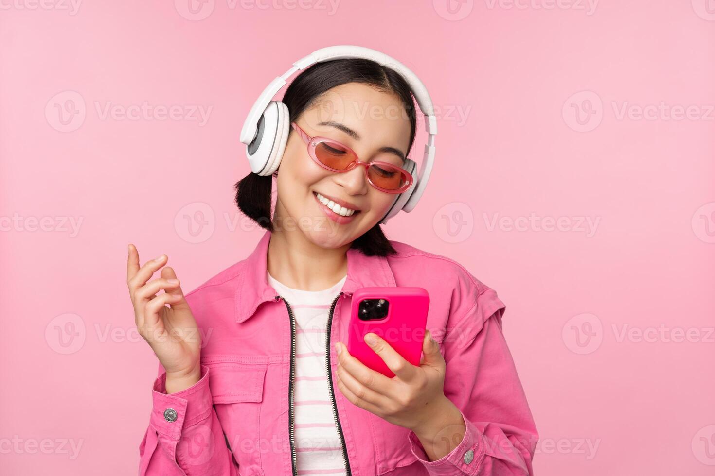 à moda ásia menina dançando com Smartphone, ouvindo música dentro fones de ouvido em Móvel telefone aplicativo, sorridente e rindo, posando contra Rosa fundo foto