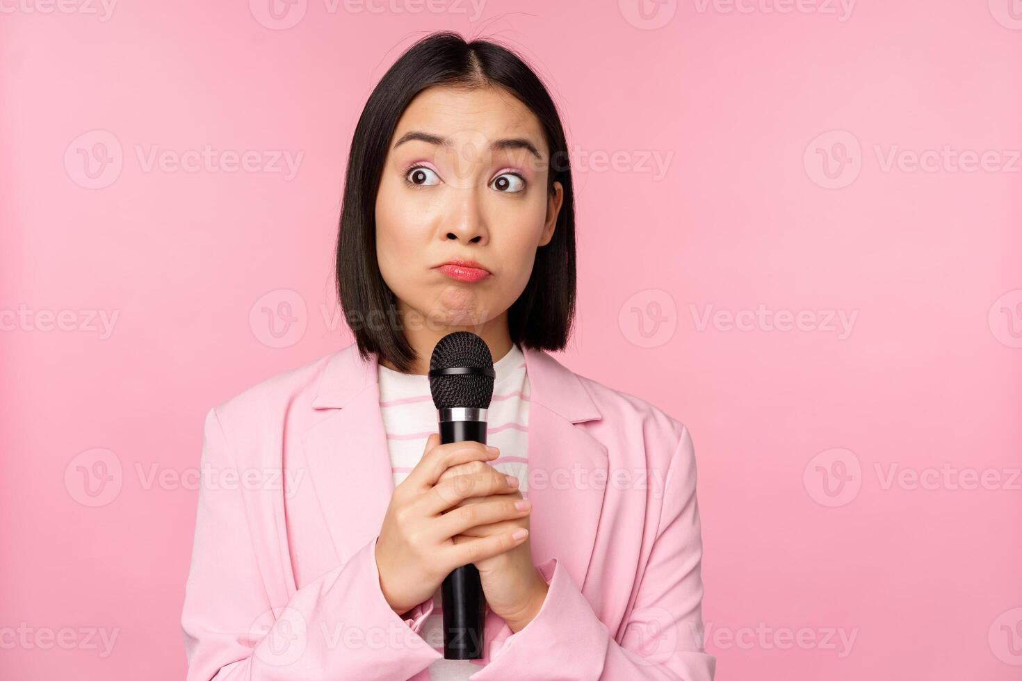 nervoso fêmea empreendedor dando discurso com microfone, segurando microfone e olhando a parte, de lado ansiosamente, posando dentro terno sobre Rosa fundo foto