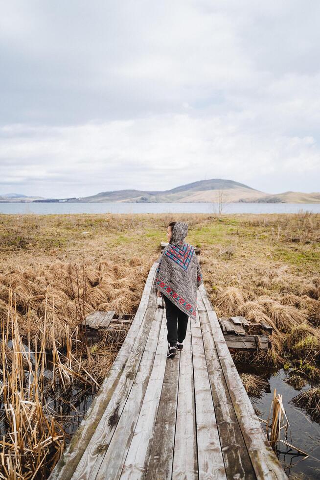 uma jovem mulher dentro uma colori poncho anda em ao longo a velho de madeira ponte através uma campo com uma pântano. a ponte vai em linha reta para a lago, atrás que a montanhas ascender. panorama em lago bannoye foto