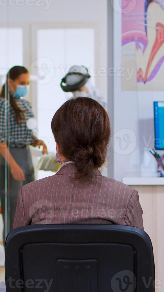 estomatológico médico com proteção terno convidativo Próximo paciente dentro dental quarto para dentes exame durante coronavírus. assistente e dentista médico vestindo geral, face escudo, mascarar, luvas foto