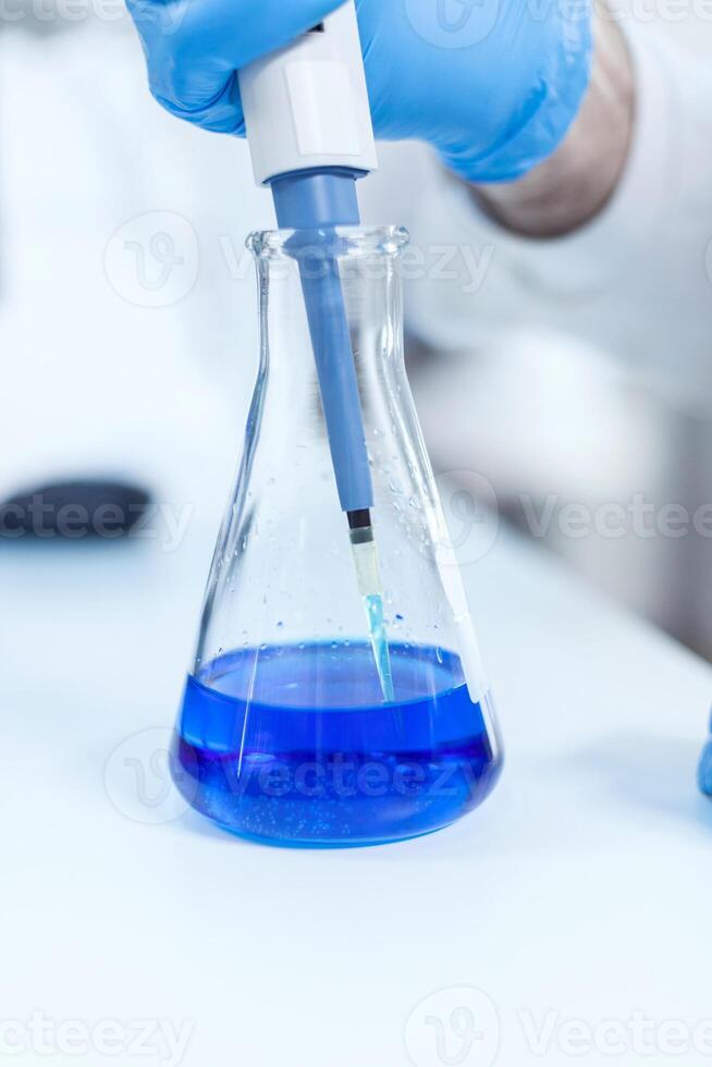 masculino técnico dentro genética laboratório usando conta-gotas para azul líquido. Senior profissional químico usando pipeta com azul solução para microbiologia testes. foto