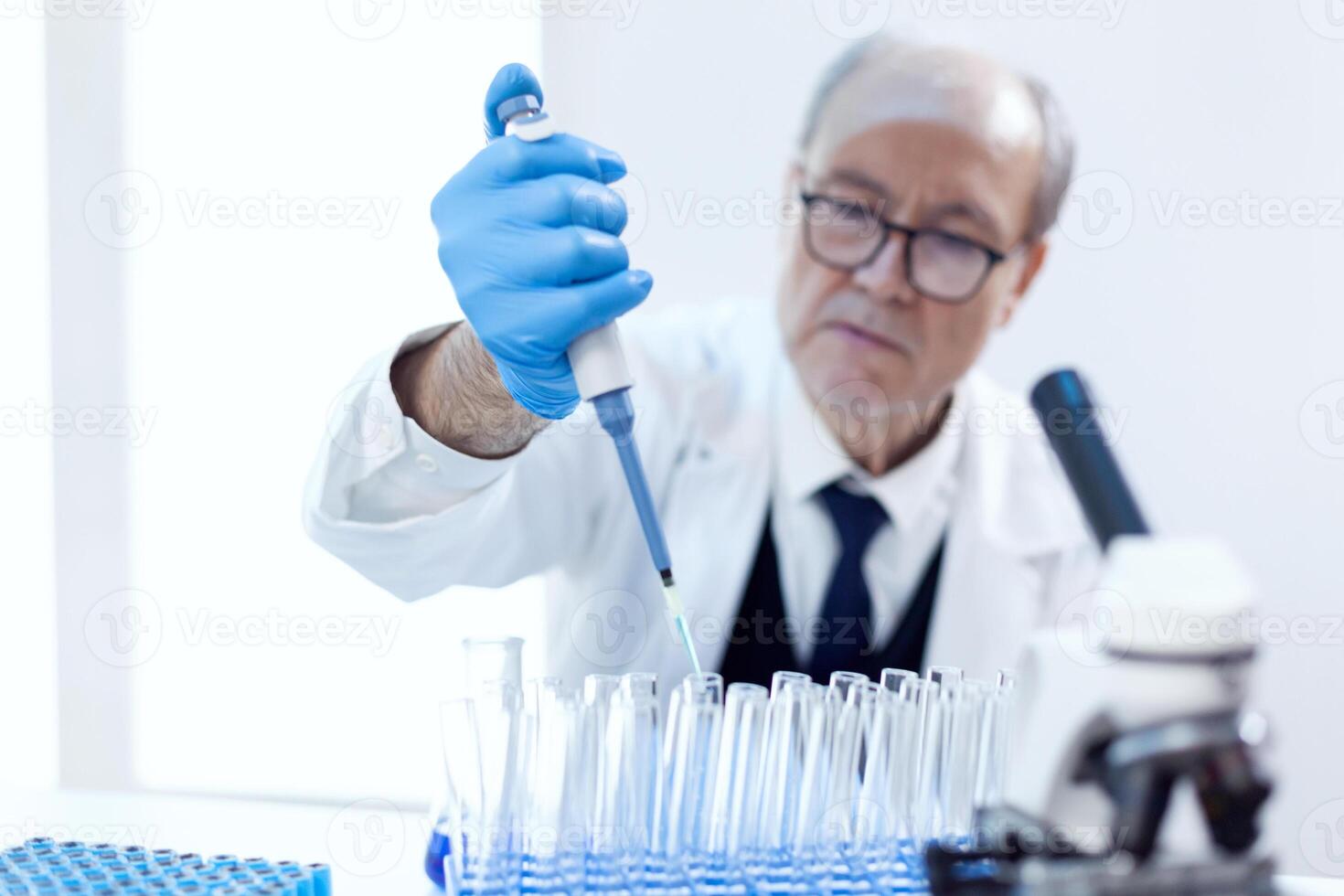farmacêutico cientista derramando azul substância dentro teste tubos sentado em dele local de trabalho com conta-gotas pipeta. Senior profissional químico usando pipeta com azul solução para microbiologia testes. foto