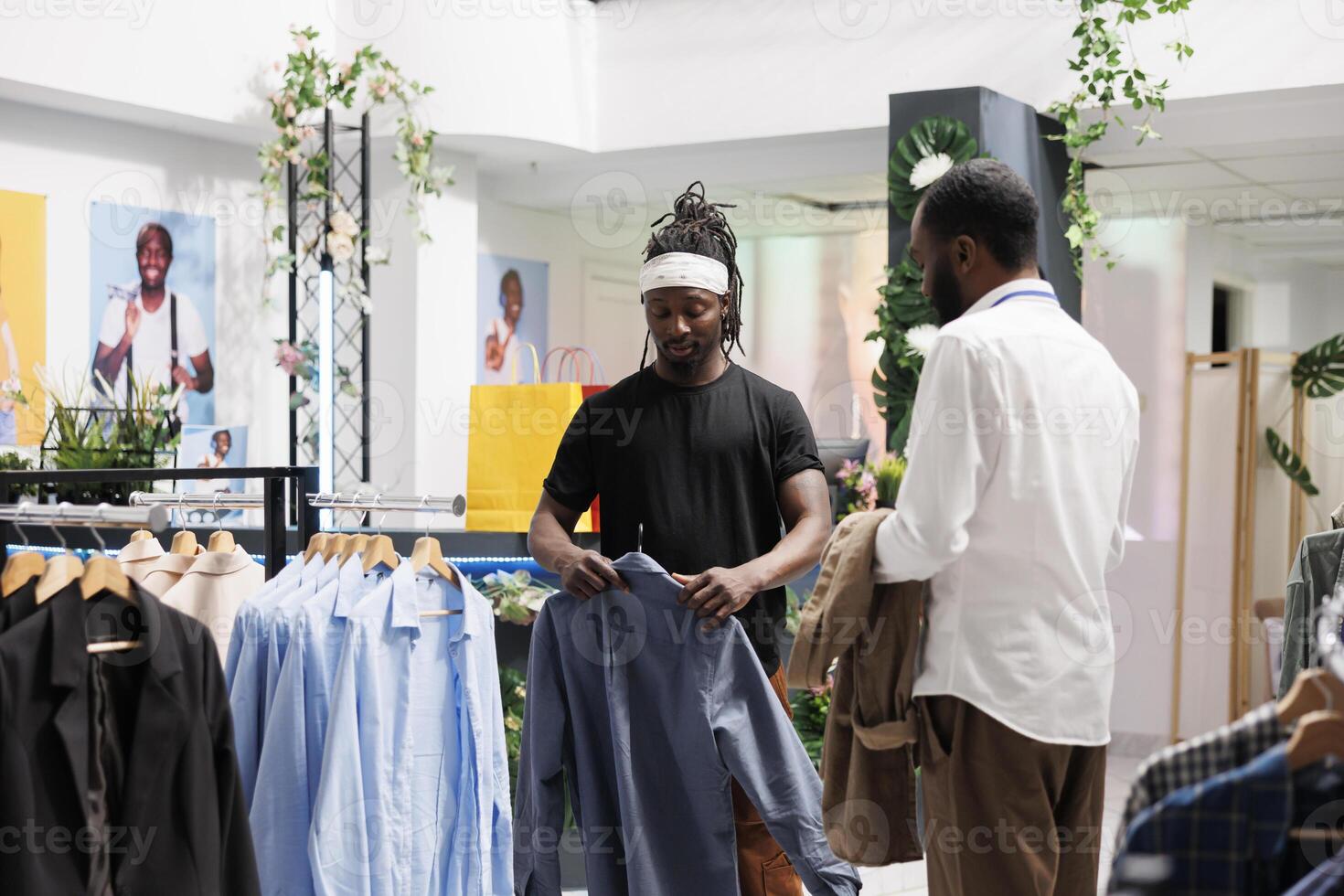 africano americano comprador verificação à moda camisa em cabide dentro roupas loja enquanto assistente dando conselho. cliente Perguntando compras Shopping empregado para Socorro enquanto escolhendo casual equipamento foto