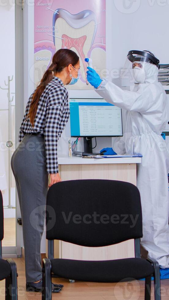 dental assistente com macacão contra coronavírus dando Formato para preencher Fora depois de medindo paciente temperatura. paciente com face proteção mascarar escrevendo em cadastro documento dentro estomatomolia clínica foto