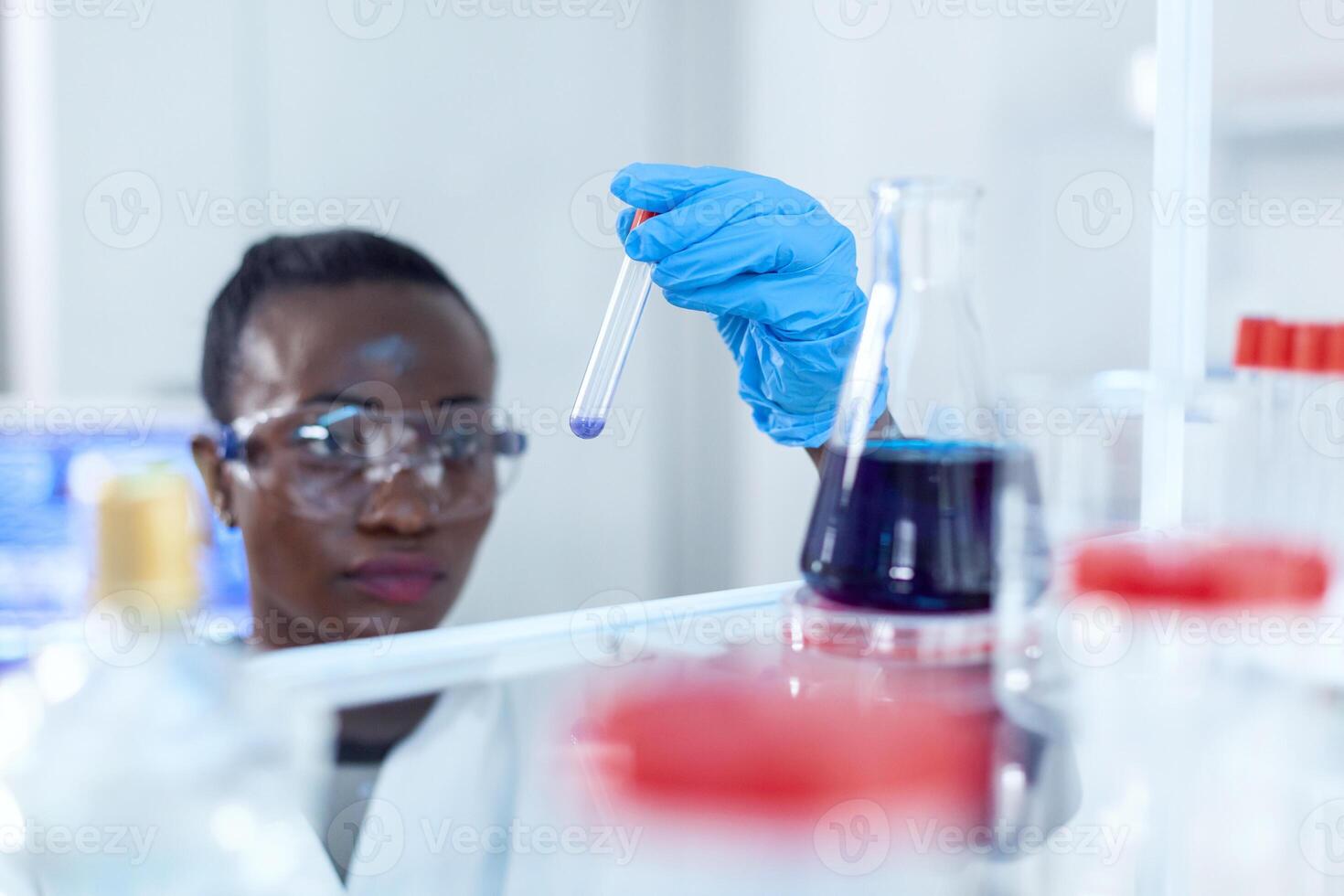 africano cientista encarando às amostra azul líquido dentro teste tubo químico. Preto investigador dentro estéril laboratório conduzindo farmacologia experimentar vestindo casaco. foto