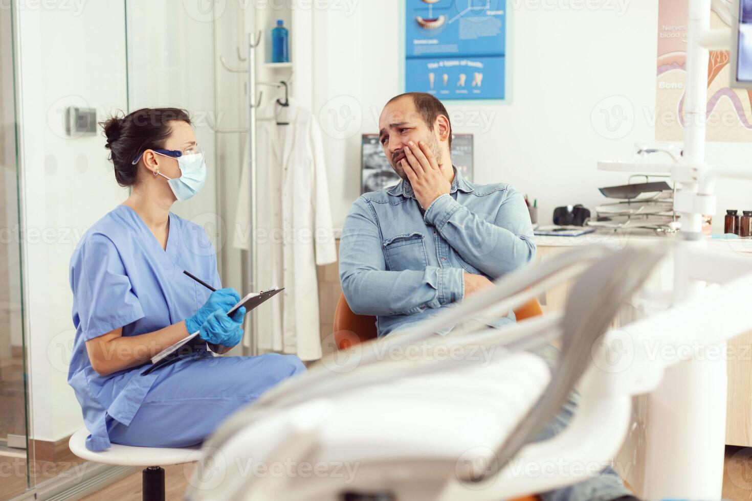 dental assistente escrevendo estomatologia tratamento em prancheta dentro dental clínica durante doente paciente Verifica acima. mulher enfermeira falando com homem sobre dentes problema esperando médico exame foto