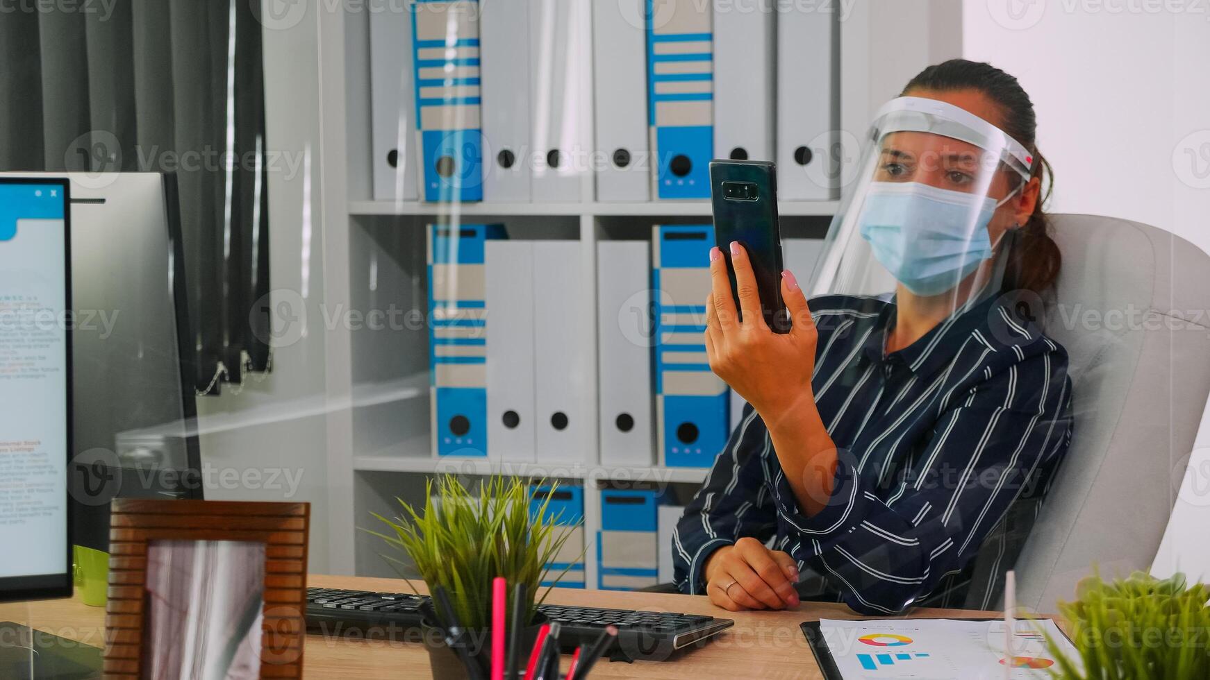 empreendedor vestindo face proteção contra coronavírus usando telefone para videoconferência falando com remotamente colega de trabalho sentado dentro Novo normal escritório corporativo prédio. mulher respeitando social distância foto