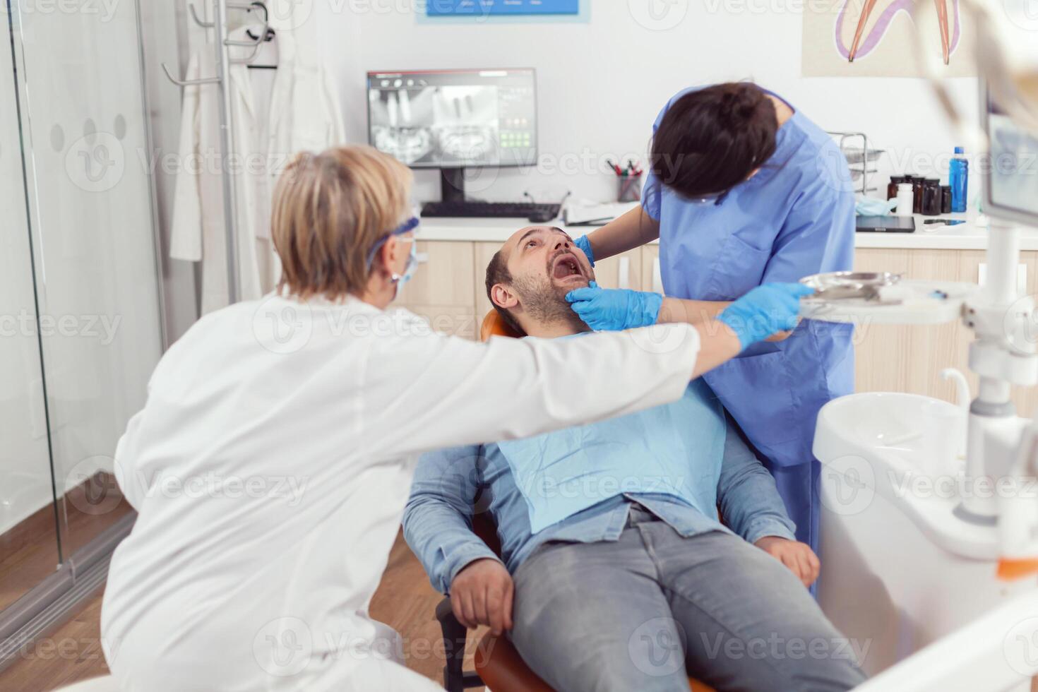 doente homem sentado em dental cadeira enquanto médico enfermeira olhando para dentro boca verificação dentes saúde durante estomatologia consulta. Senior dentista mulher preparando Ferramentas para oral cirurgia foto