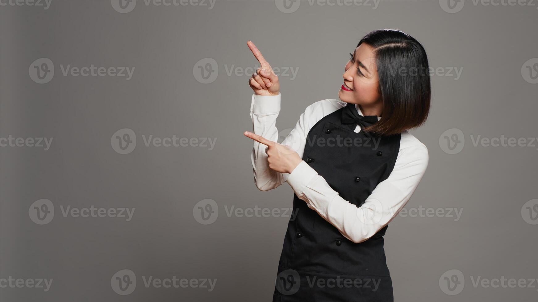 ásia garçonete apontando acima para a propaganda dentro estúdio, indicando direção acima dela cabeça para crio promocional de Anúncios. mulher refeições empregado pontos às alguma coisa para cima. Câmera uma. foto