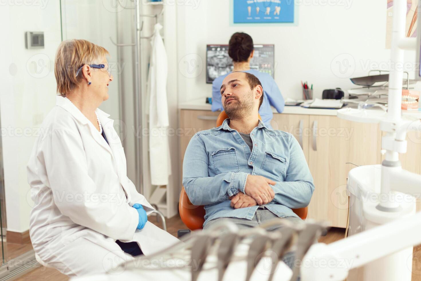 Senior dentista mulher discutindo com paciente sobre dor de dente tratamento durante estomatológico consulta dentro dental hospital escritório. doente homem sentado em dental cadeira esperando dente radiografia foto