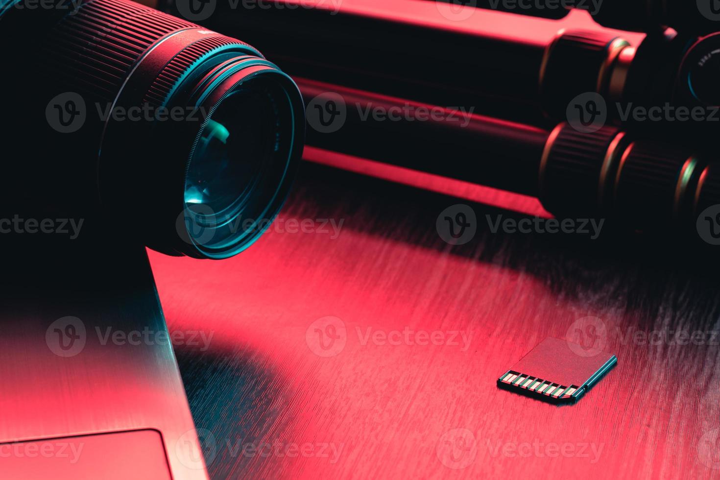 lente da câmera, cartão, laptop e tripé na mesa de madeira. área de trabalho da mesa. luz vermelha e azul foto