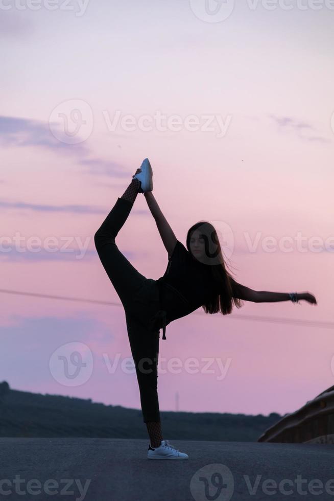 linda jovem morena posando em uma estrada no meio do campo ao pôr do sol, balé, silhueta foto