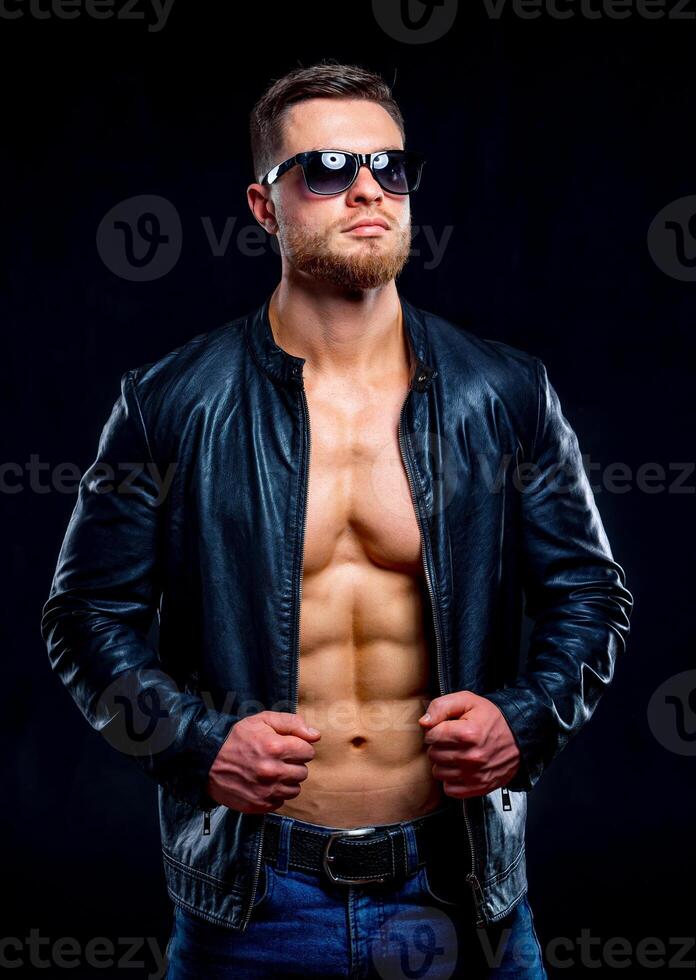 macho homem com perfeito corpo dentro oculos de sol com aberto couro Jaqueta revelador muscular peito e tronco. fechar-se foto