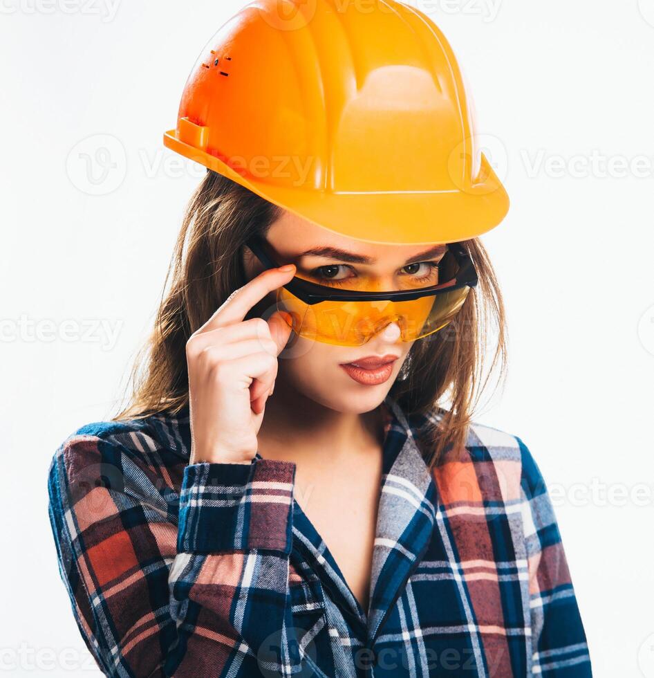 feliz jovem mulher é vestindo laranja segurança capacete e xadrez camisa olhando através amarelo segurança óculos. isoalted fundo. foto