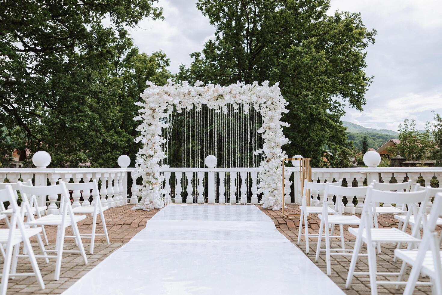 Casamento arco fez do branco flores em uma fundo do árvores uma branco caminho para a arco, muitos branco cadeiras. preparação para a Casamento cerimônia foto