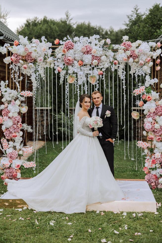 uma bonito noivo abraços dele noiva dentro uma exuberante branco vestir e sorrisos dentro uma lindo ao ar livre contexto. debaixo a aberto céu. Alto qualidade foto. uma recém-casado casal poses juntos em uma ensolarado verão dia. foto