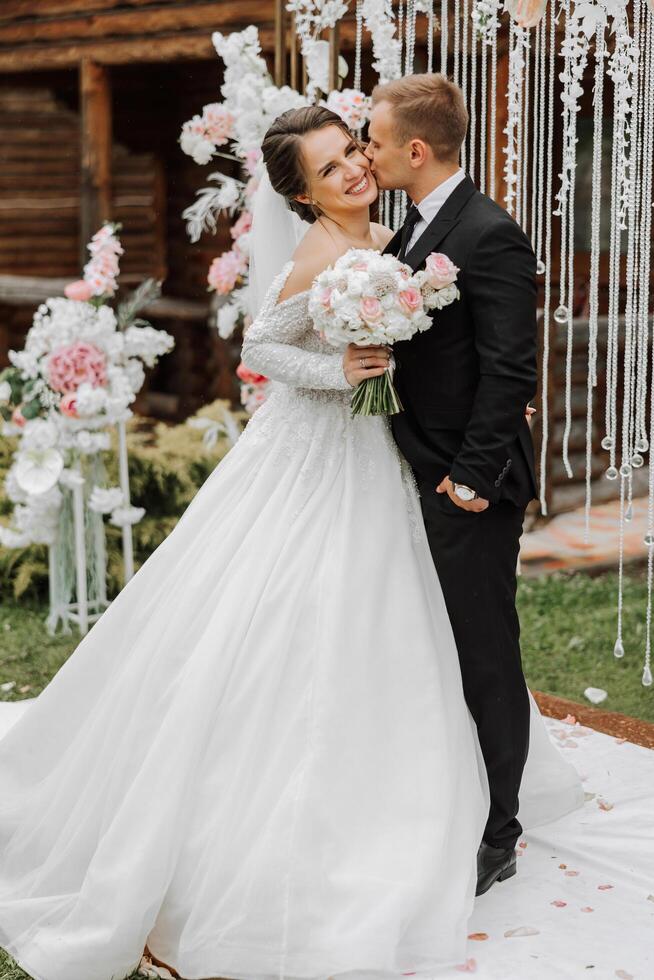 uma bonito noivo abraços dele noiva dentro uma exuberante branco vestir e sorrisos dentro uma lindo ao ar livre contexto. debaixo a aberto céu. Alto qualidade foto. uma recém-casado casal poses juntos em uma ensolarado verão dia. foto