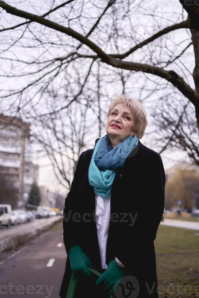 retrato do uma à moda velho mulher dentro uma Preto casaco com verde acessórios em uma Primavera rua foto