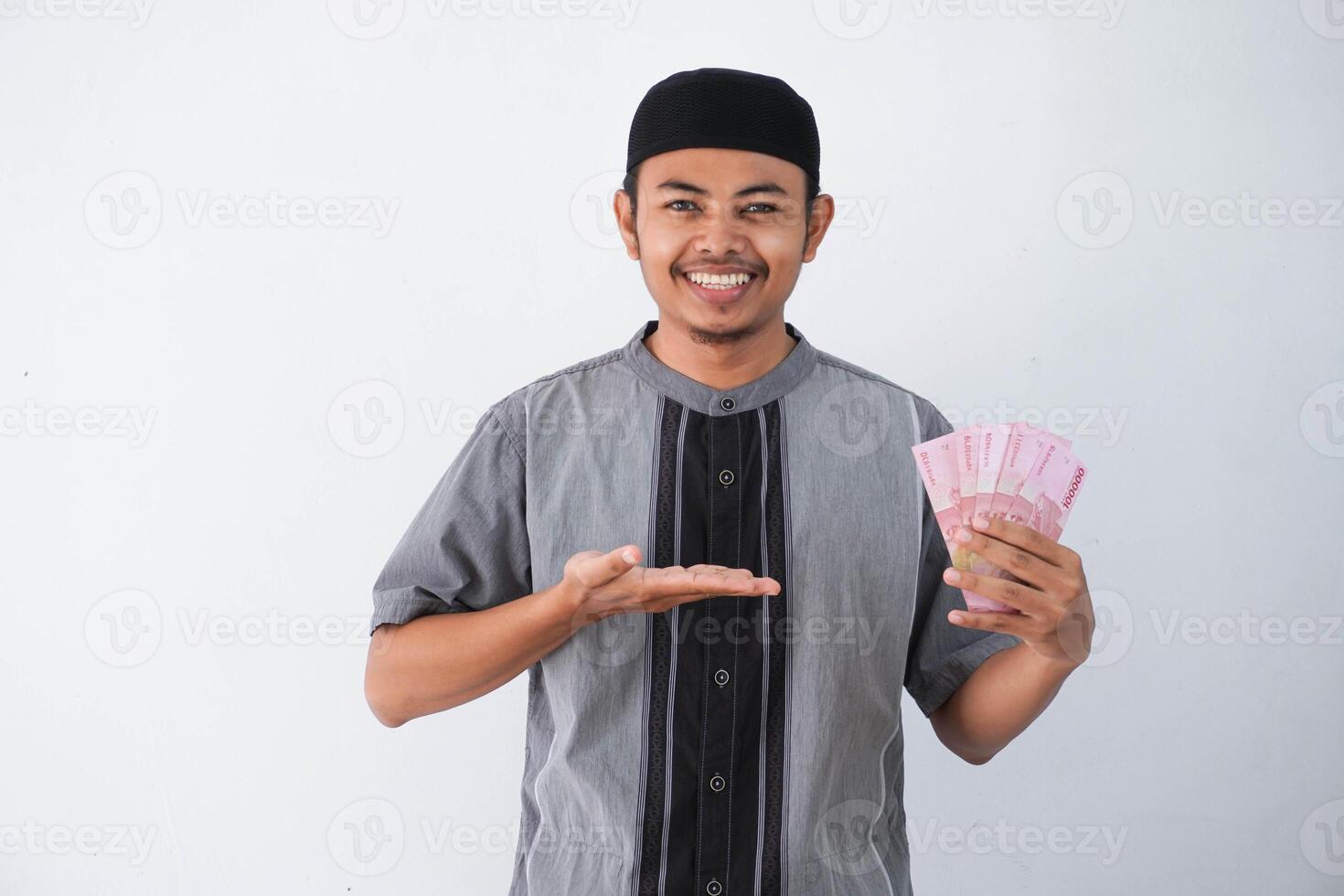 sorridente ou feliz jovem ásia muçulmano homem segurando e apontando papel dinheiro presente thr a partir de família Ramadã eid al-fitr Tempo vestindo cinzento muçulmano roupas isolado em branco fundo foto