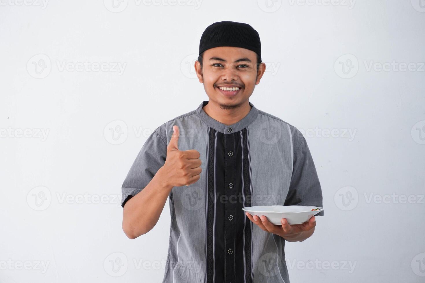 sorridente ou feliz jovem ásia muçulmano homem mostrando animado expressão dando polegar acima enquanto segurando esvaziar jantar prato vestindo cinzento muçulmano roupas isolado em branco fundo foto
