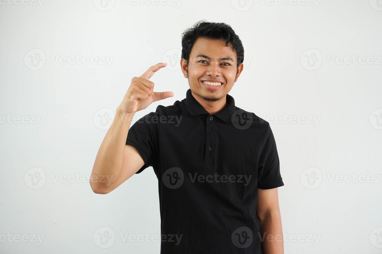 jovem ásia homem posando em uma branco pano de fundo segurando alguma coisa pequeno com dedos indicadores, sorridente e confiante, vestindo Preto pólo t camisa. foto