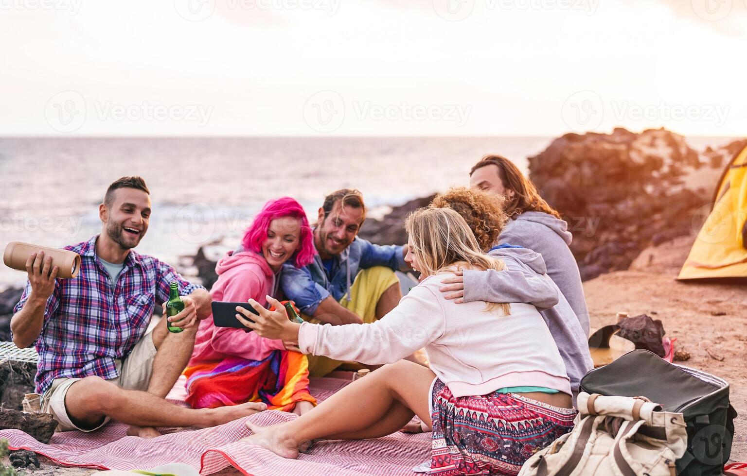 feliz amigos fazendo festa às pôr do sol enquanto acampamento Próximo a de praia - milenar jovem pessoas tendo Diversão bebendo Cerveja e usando Móvel Smartphone ao ar livre - verão, férias, tecnologia e juventude conceito foto