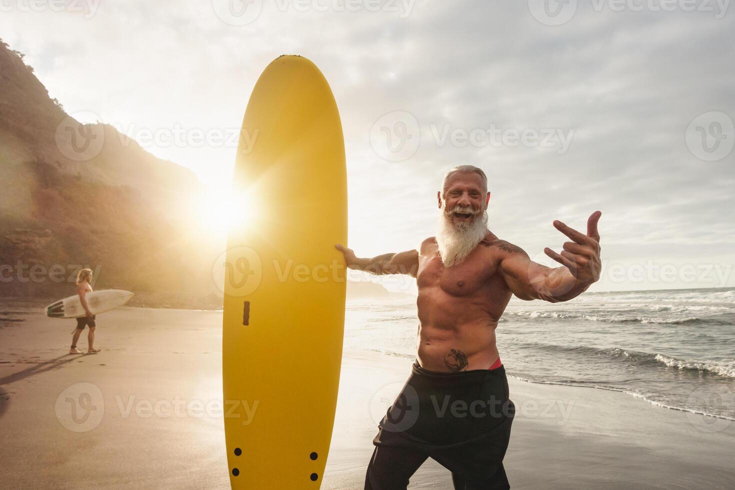 feliz em forma Senior tendo Diversão surfar às pôr do sol Tempo - desportivo barbudo homem Treinamento com prancha de surfe em a de praia - idosos saudável pessoas estilo de vida e extremo esporte conceito foto