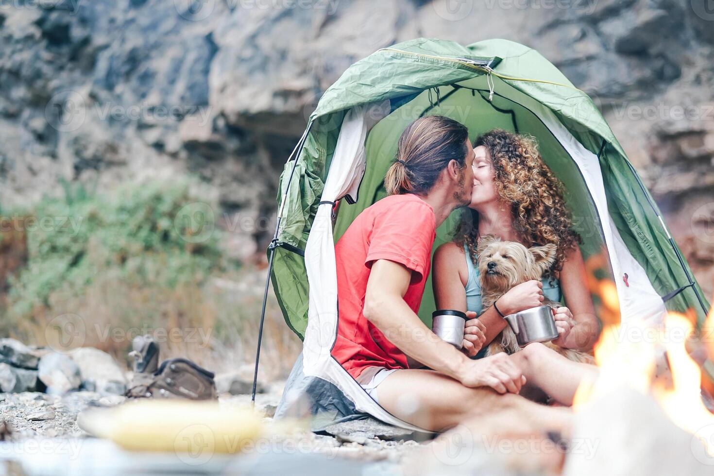 viagem casal acampamento dentro Rocha montanhas depois de uma caminhada dia - feliz pessoas bebendo quente chá dentro barraca com seus cachorro Próximo para fogueira - esporte, relação, amor e período de férias conceito foto