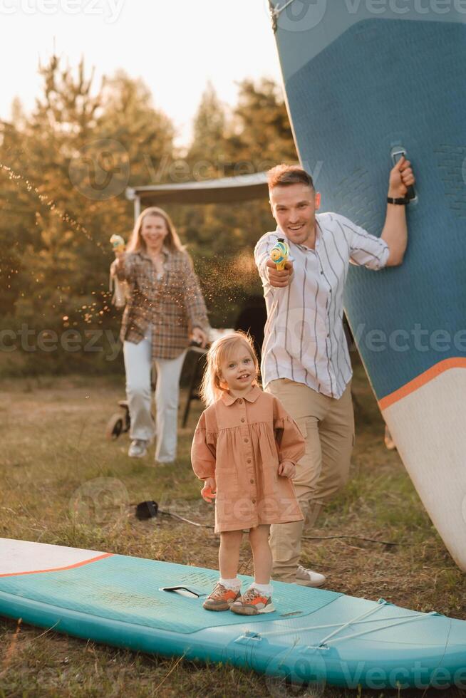 a família é em repouso Próximo para seus Móvel lar. pai, mãe e filha jogar em sup Pranchas com água pistolas perto a motorhome foto