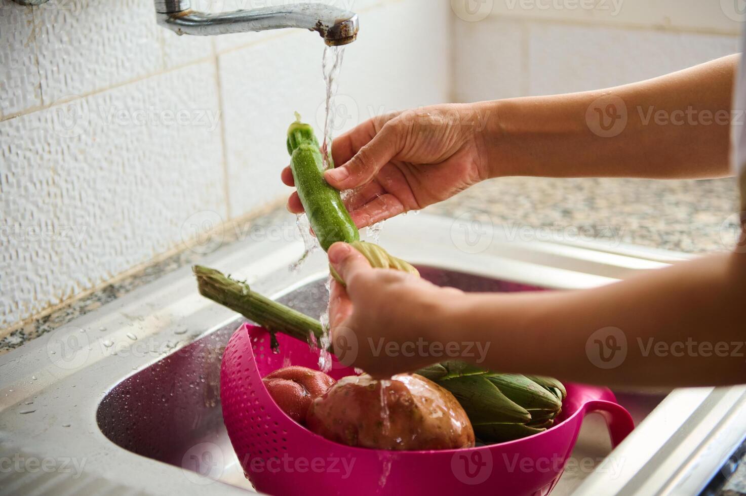 fechar-se do fêmea mãos lavando abobrinha e legumes debaixo fluindo água, em pé de uma inoxidável Pia dentro casa cozinha foto