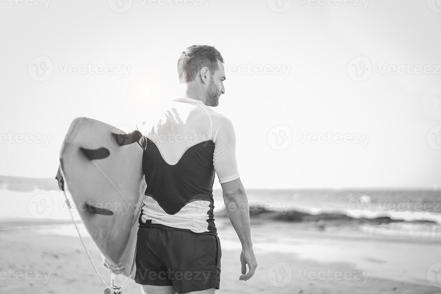 jovem surfista segurando dele prancha de surfe em a de praia - bonito homem esperando ondas para surfar - Preto e branco edição - pessoas, esporte e estilo de vida conceito foto
