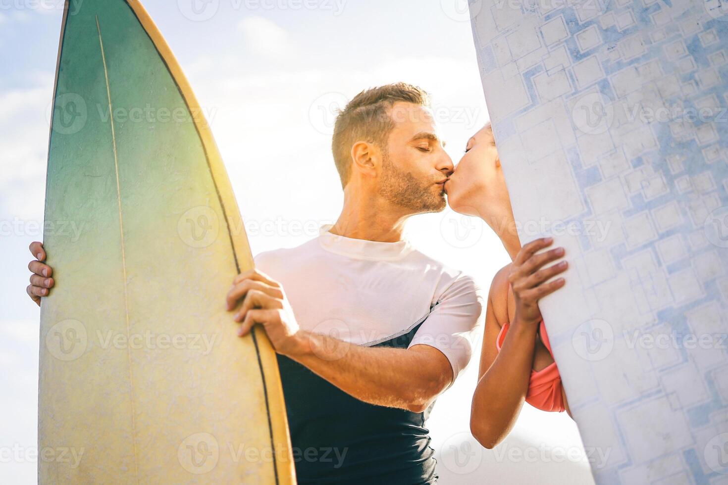 saúde jovem casal do surfistas se beijando às pôr do sol em a de praia segurando pranchas de surf - feliz amantes tendo uma concurso momento com uma beijo enquanto surfar juntos - pessoas, amor, esporte e estilo de vida conceito foto