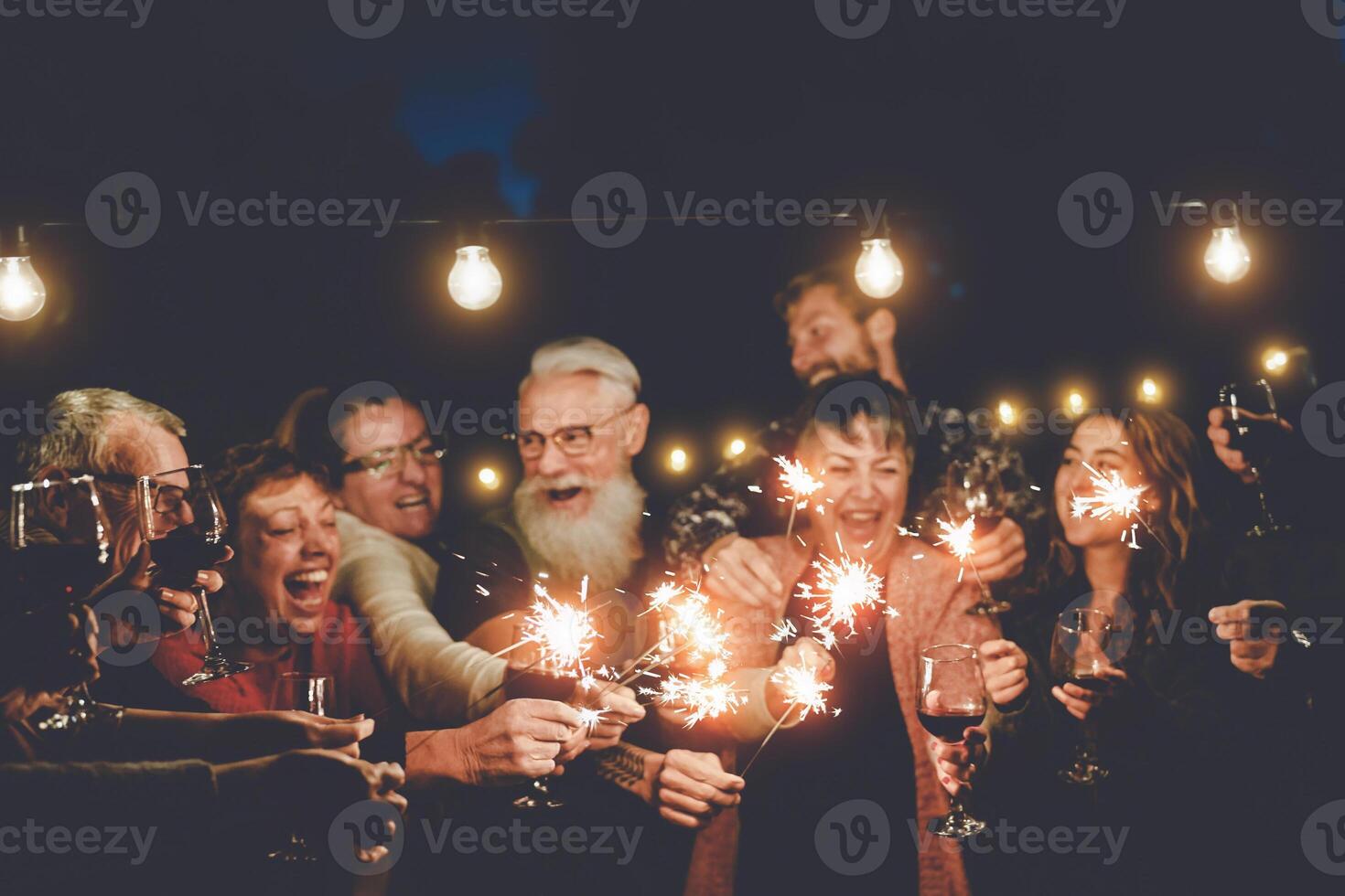 feliz família tendo às jantar festa ao ar livre - grupo do multirracial Mais velho e jovem pessoas a comemorar juntos bebendo vinho segurando fogos de artifício estrelinhas - conceito do juventude e idosos paternidade foto