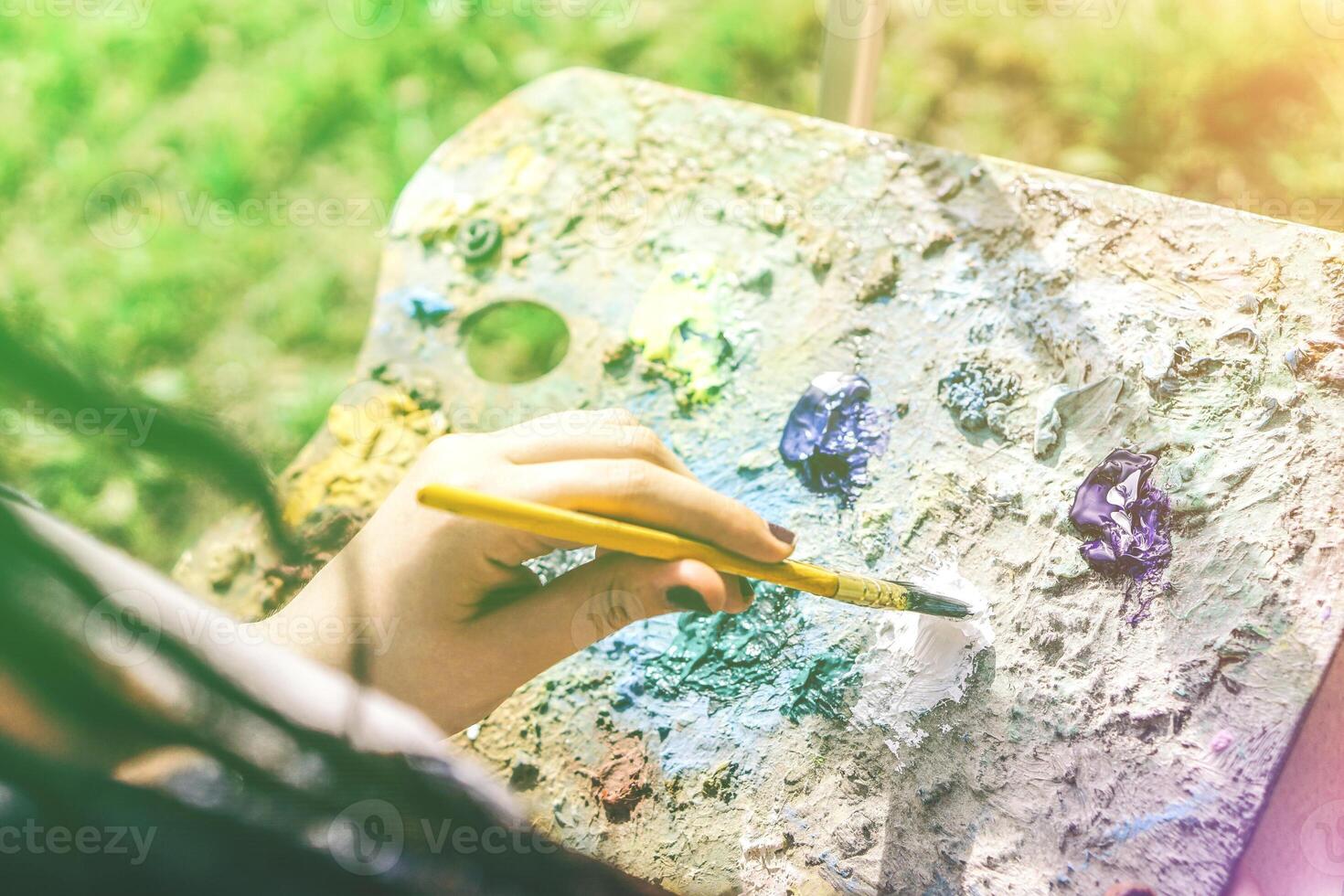 jovem artista pintura dentro uma parque ao ar livre - fechar acima do pintor com tranças Penteado trabalhando em dela arte dentro uma colorida jardim - conceito do pessoas expressando artes - foco em mão pintura foto